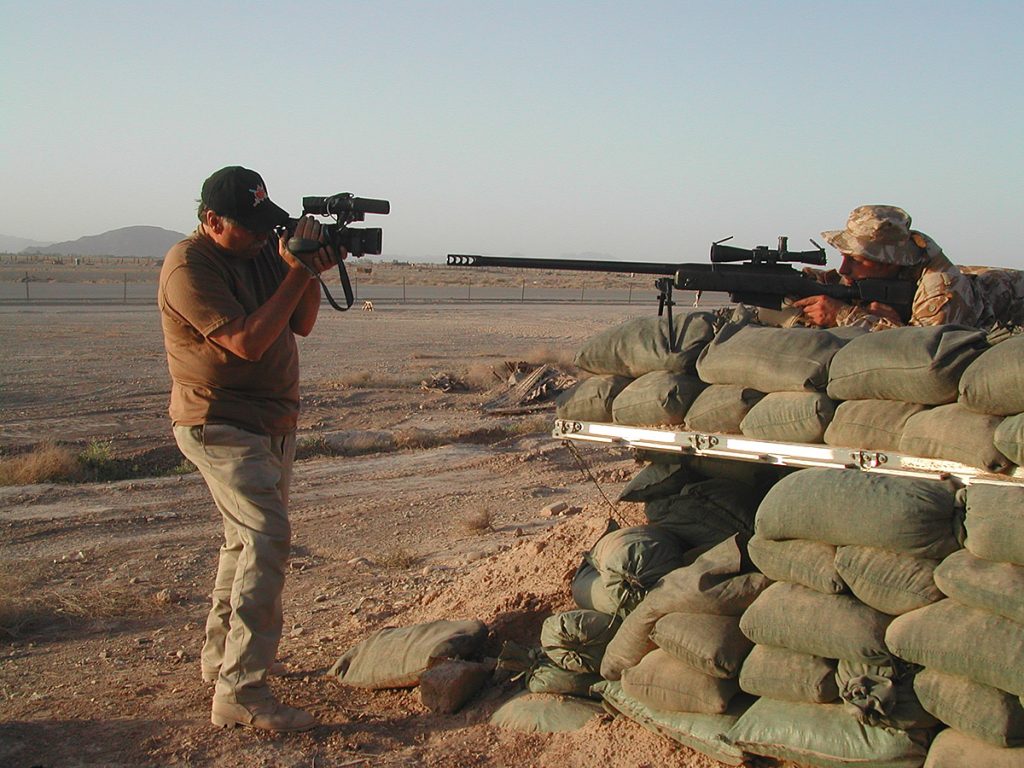 Un homme en train de filmer un soldat pointant un fusil derrière un tas de sacs de sable dans le désert