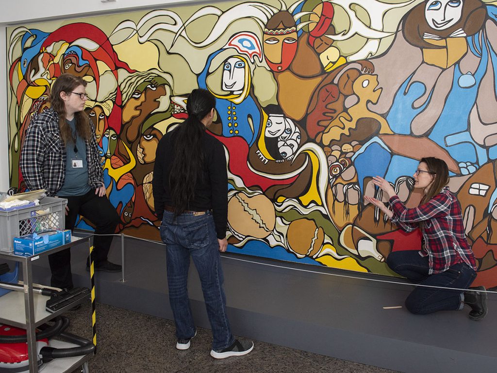 Une femme accroupie parle à deux hommes debout en montrant une murale colorée