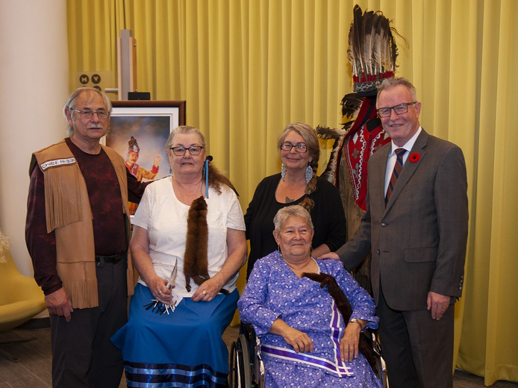 Groupe de cinq personnes, dont trois debout et deux assises, notamment une femme en fauteuil roulant, devant une image encadrée et un mannequin vêtu d’une tenue cérémonielle autochtone