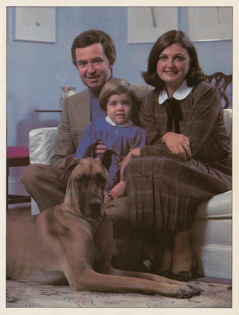 Photo de deux adultes et un enfant habillés formellement assis sur un divan avec un chien à leurs pieds.