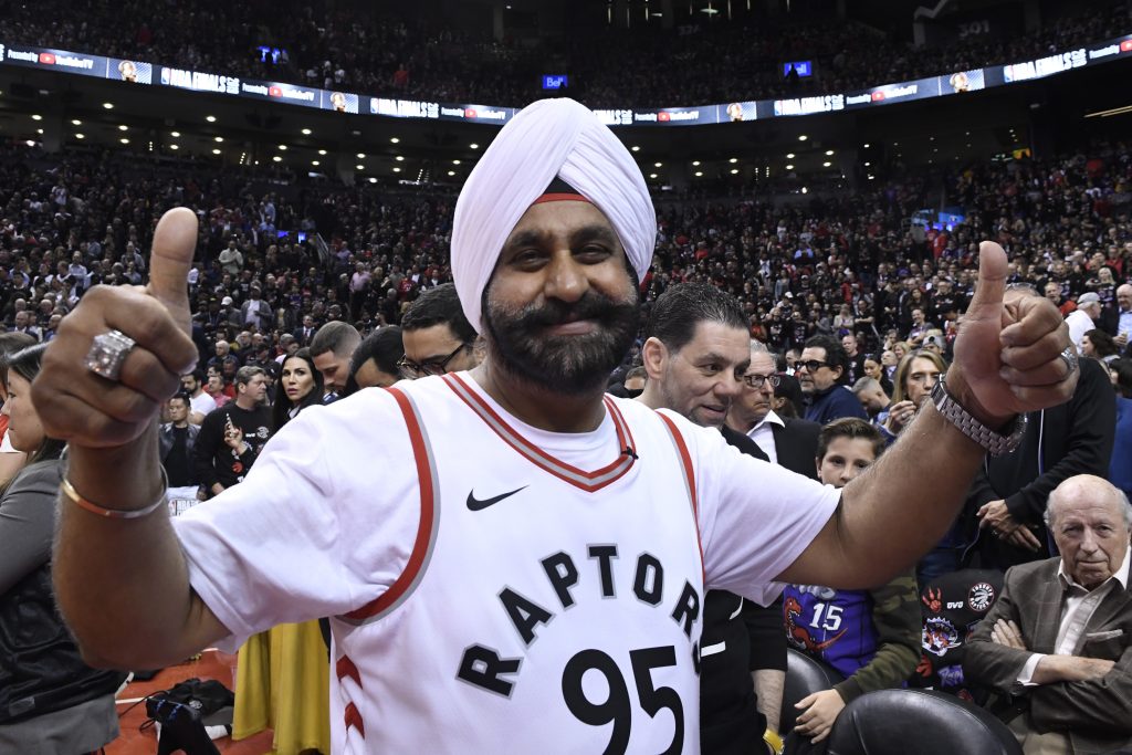 Un homme à la peau foncée et à la barbe noire porte un turban blanc et un gilet de sport blanc portant l’inscription « Raptors 95 », tout en levant deux pouces et en souriant à l’appareil photo. À l’arrière-plan, on aperçoit un grand stade rempli de gens.