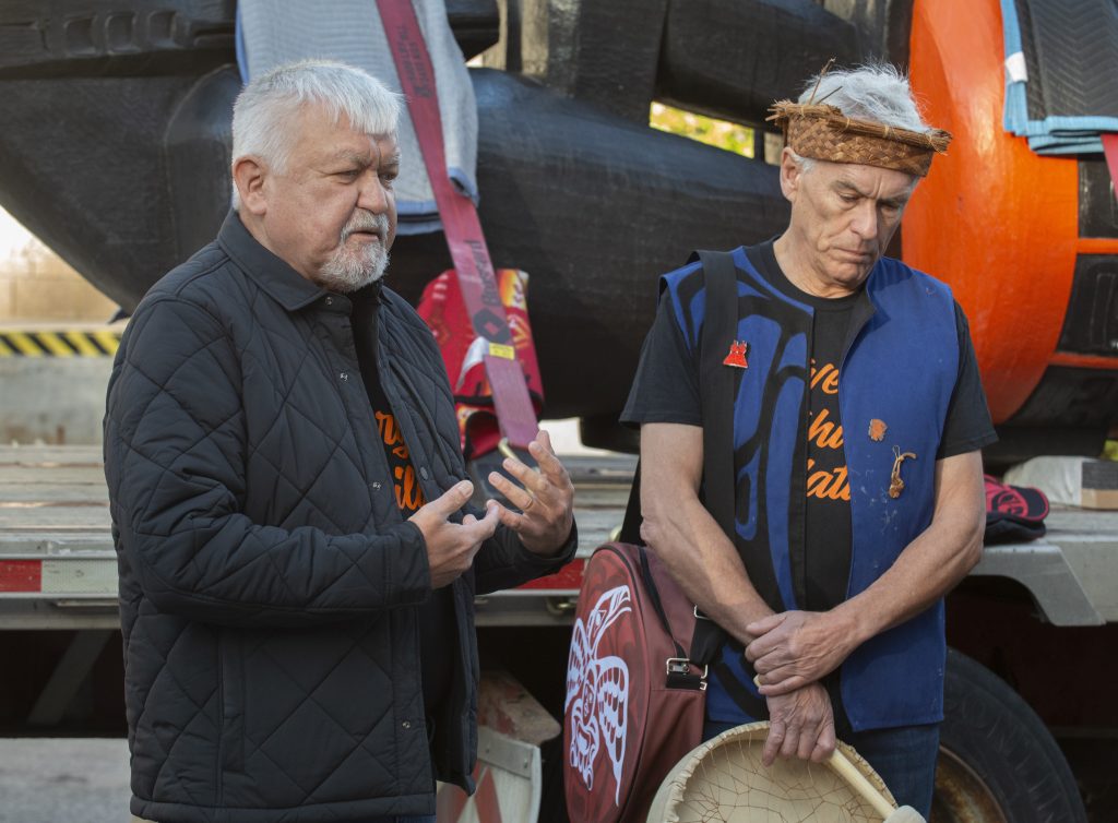 Deux hommes à la peau basanée et aux cheveux blancs se tiennent devant une grande sculpture orange et noir.