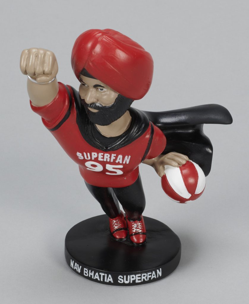 Une figurine en plastique d’un homme à la peau foncée et à la barbe noire portant un turban rouge, une chemise rouge sur laquelle on peut lire l’inscription « Superfan 95 » et une cape noire, et tenant un ballon de basket rouge et blanc.