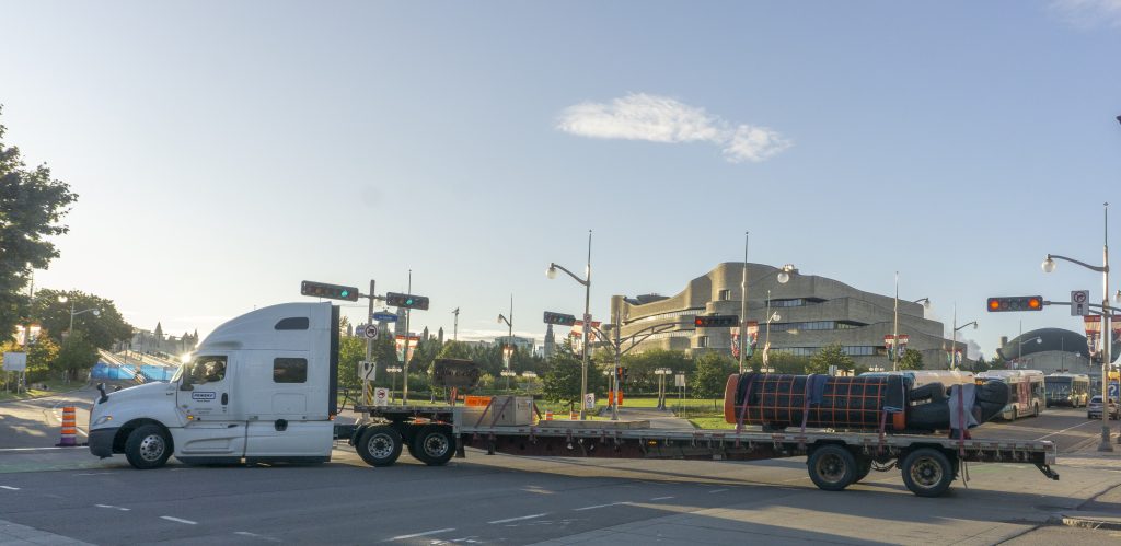 Un camion de transport passe devant la façade du Musée, trainant le Monument allongé sur une longue remorque.