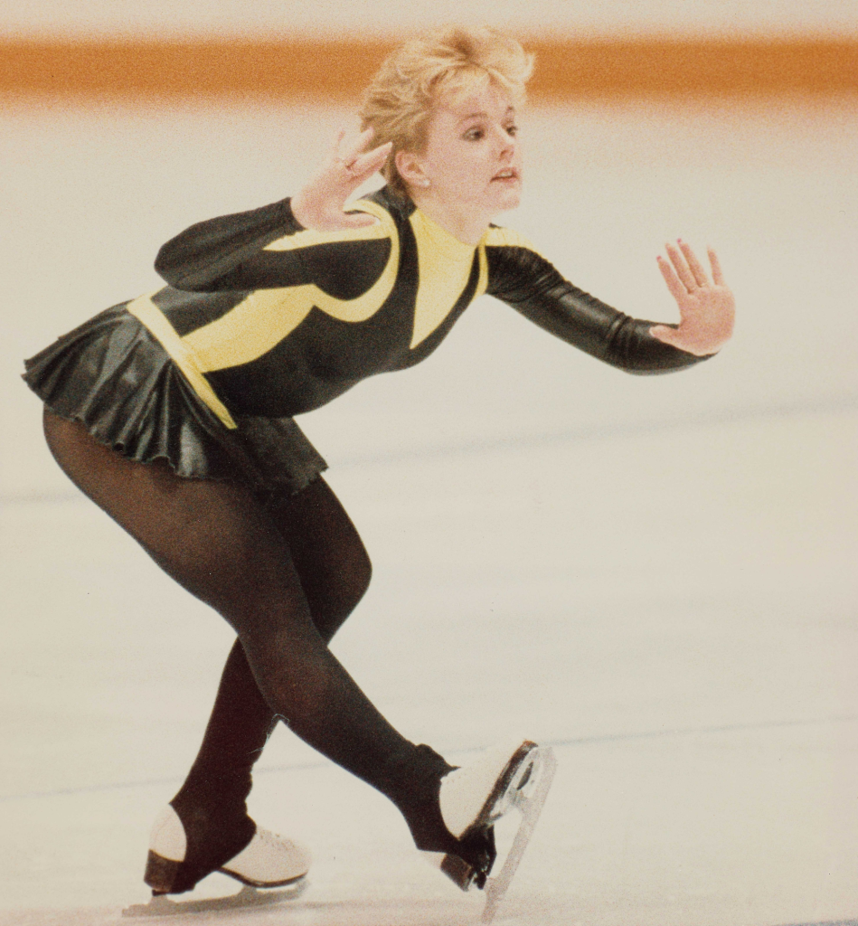 Une femme blonde vêtue d’un justaucorps jaune et noir et d’une jupe courte patine en se penchant en avant et en faisant des gestes avec les mains.