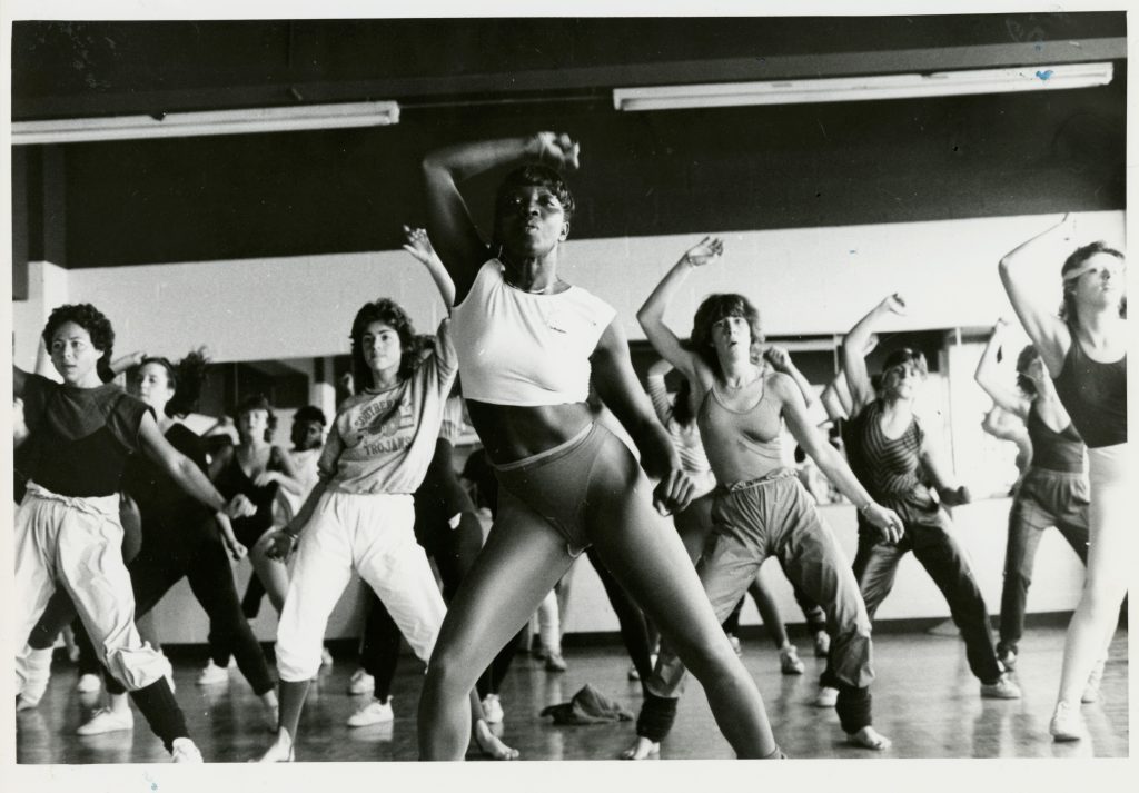 Une salle de cours de conditionnement physique bondée de personnes portant divers vêtements de sport. Le cours est donné par une femme noire athlétique vêtue d’un haut court et de jambières.