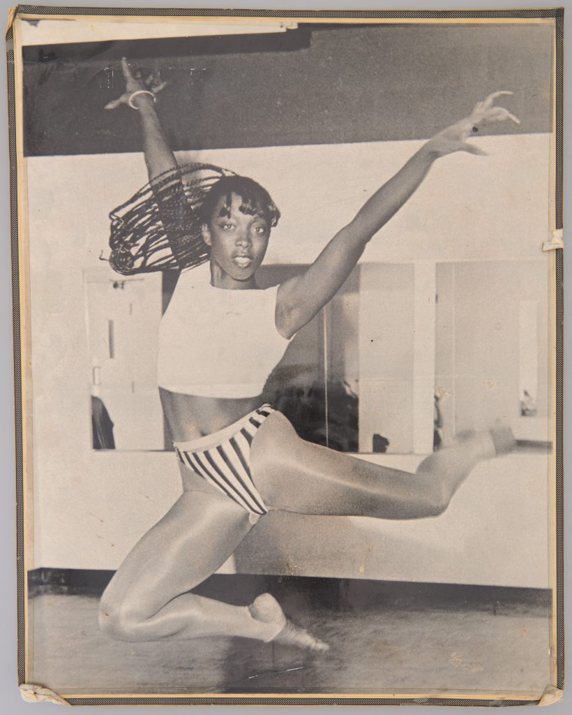 Une femme noire athlétique saute dans les airs, les bras écartés. Elle a de longues tresses qui volent sur le côté et porte un haut court et des jambières de danse. 