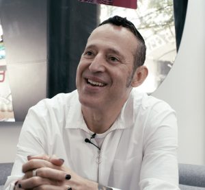 A man in a white shirt holds his hands together and smiles.