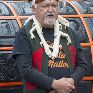 Un homme au teint moyen portant un couvre-chef en cuir et en fourrure, un collier de fourrure et un gilet en tissu couvert de motifs simples et colorés devant une très grande surface sculptée orange et noire.