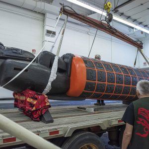 Un palan est fixé au monument, qui est placé sur une plateforme de transport. Le monument est essentiellement cylindrique, peint en noir avec un motif de grille orange, et un très grand corbeau regardant vers le bas est assis au sommet du cylindre.