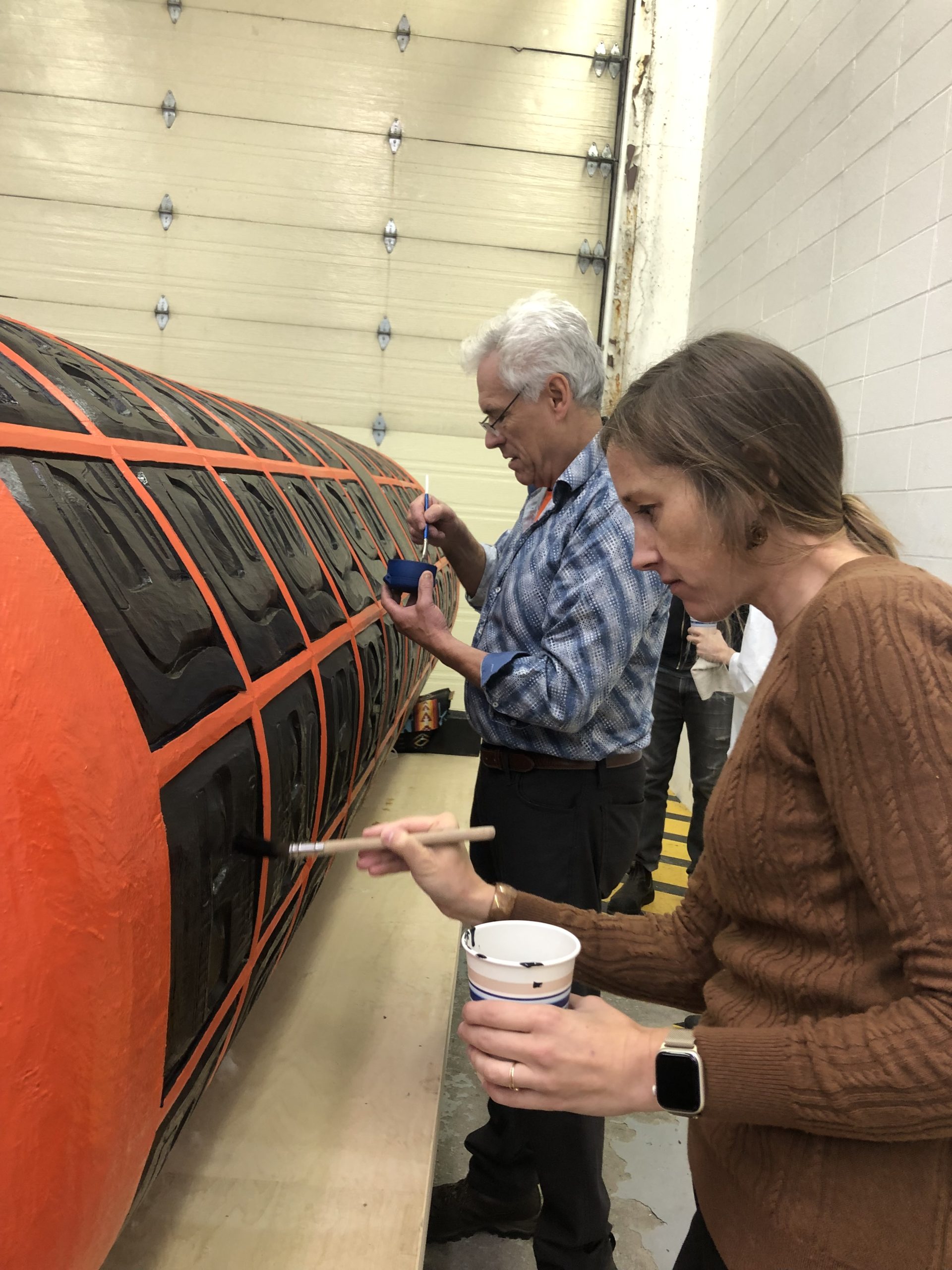 few people stand next to a large, horizontal, cylindrical carving. They are applying paint with small brushes.