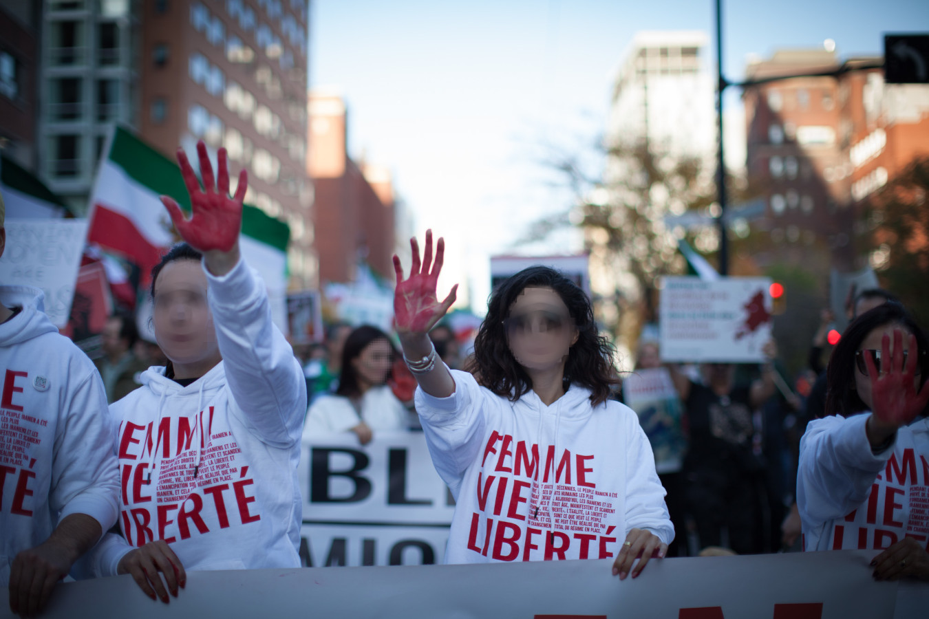 Plusieurs personnes tiennent une banderole d’une main et tendent l’autre vers l’appareil photo, couverte de peinture rouge. Elles portent des chemises sur lesquelles on peut lire « Femme Vie Liberté ».