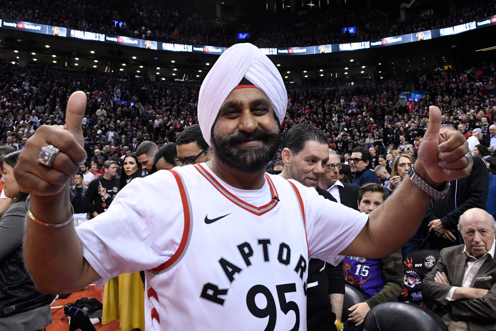 Un homme à la peau foncée et à la barbe noire porte un turban blanc et un gilet de sport blanc portant l’inscription « Raptors 95 », tout en levant deux pouces et en souriant à l’appareil photo. À l’arrière-plan, on aperçoit un grand stade rempli de gens.