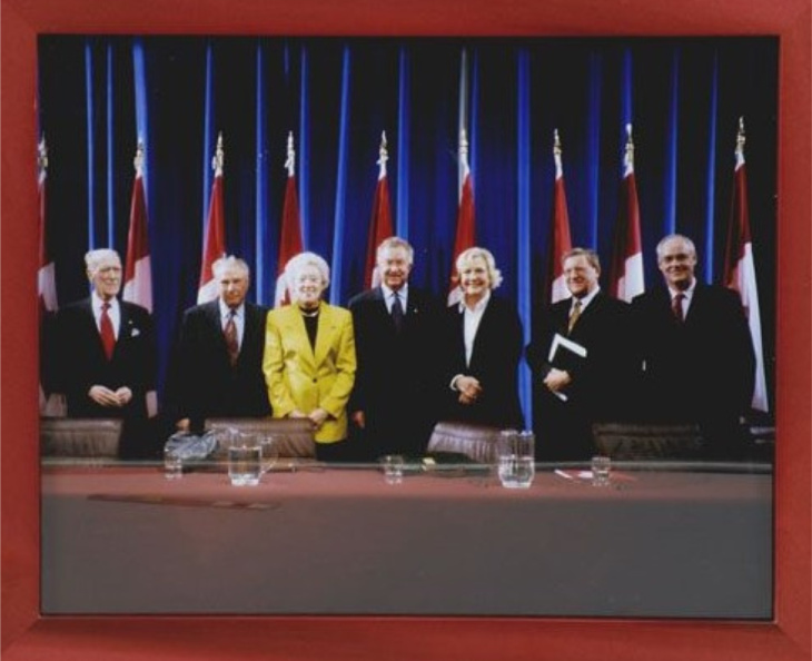 Cinq hommes et 2 femmes en tenue de travail debout derrière une table, avec des drapeaux du Canada derrière eux.