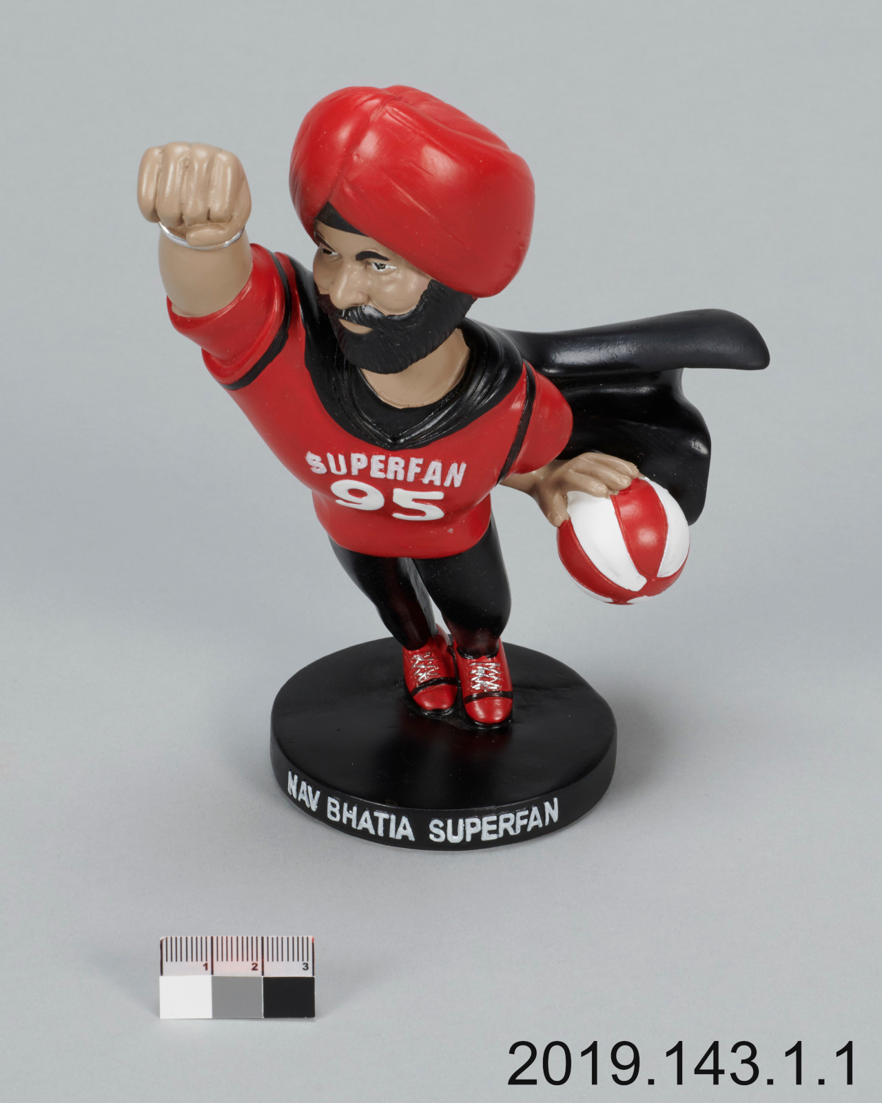 Une figurine en plastique d’un homme à la peau foncée et à la barbe noire portant un turban rouge, une chemise rouge sur laquelle on peut lire l’inscription « Superfan 95 » et une cape noire, et tenant un ballon de basket rouge et blanc.