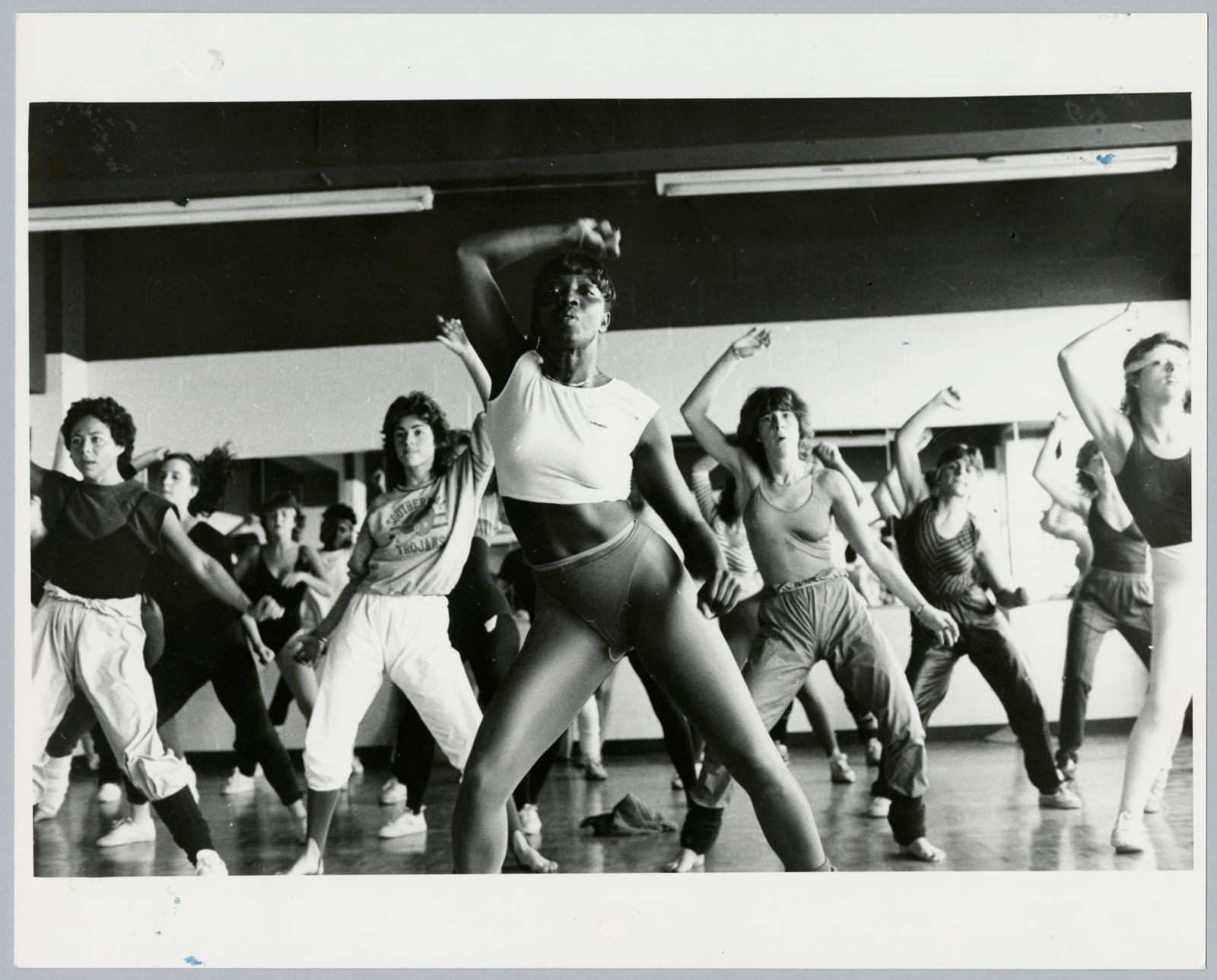 Une salle de cours de conditionnement physique bondée de personnes portant divers vêtements de sport. Le cours est donné par une femme noire athlétique vêtue d’un haut court et de jambières.