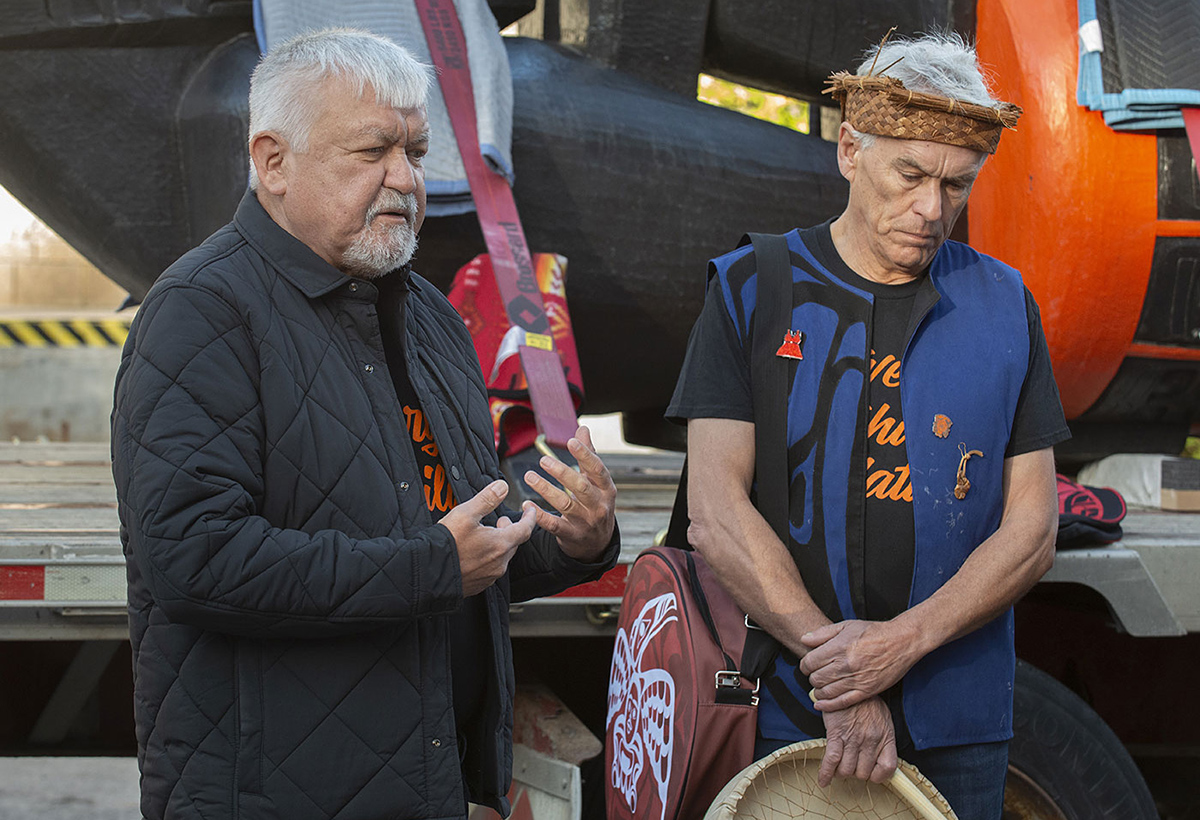 Deux hommes au teint moyen et aux cheveux blancs devant une grande sculpture orange et noire.