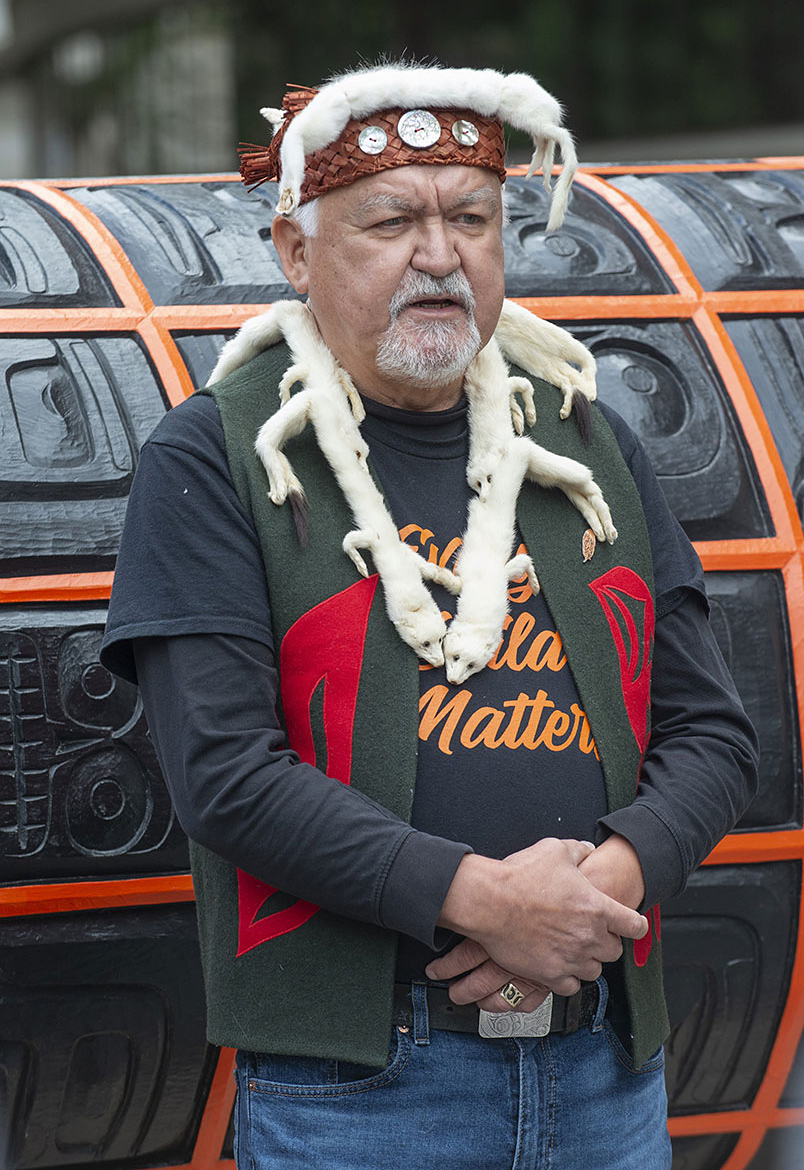 Un homme au teint moyen portant un couvre-chef en cuir et en fourrure, un collier de fourrure et un gilet en tissu couvert de motifs simples et colorés devant une très grande surface sculptée orange et noire.