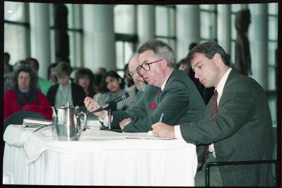 (G) Deux hommes en tenue de travail assis à une table signent des documents avec des gens assis derrière eux. (D) Document avec le titre « Bâtir ensemble l’avenir du Canada, »