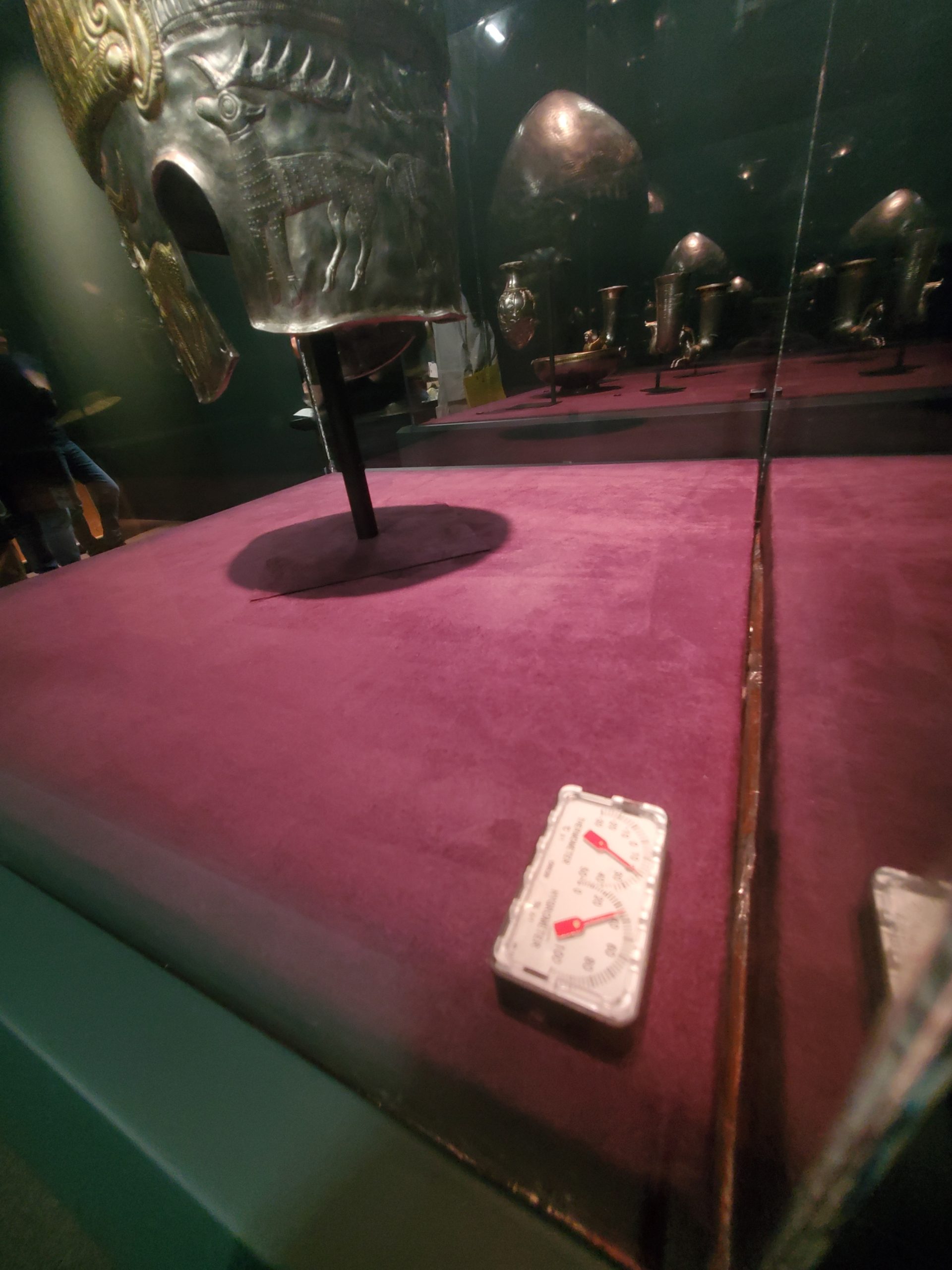 A glass display case containing an old metal helmet and a small white plastic device with two dial indicators.